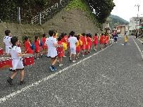 児童たちがお揃いの衣装で楽器を持って歩道を歩いている写真