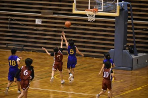 屋内のバスケットボールコートで、ユニフォームを着た児童たちがでバスケットボールに手を伸ばしている写真