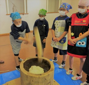 複数の児童がエプロンと三角巾をつけ、臼と杵を使って餅をついている写真