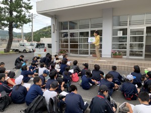 学校の校舎の前で多くの児童たちが地面に座り前に立っている人が紙を持って説明をしている写真