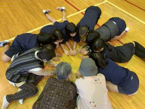床に児童と高齢者が円形に座り、全員が床に手を伸ばしている写真