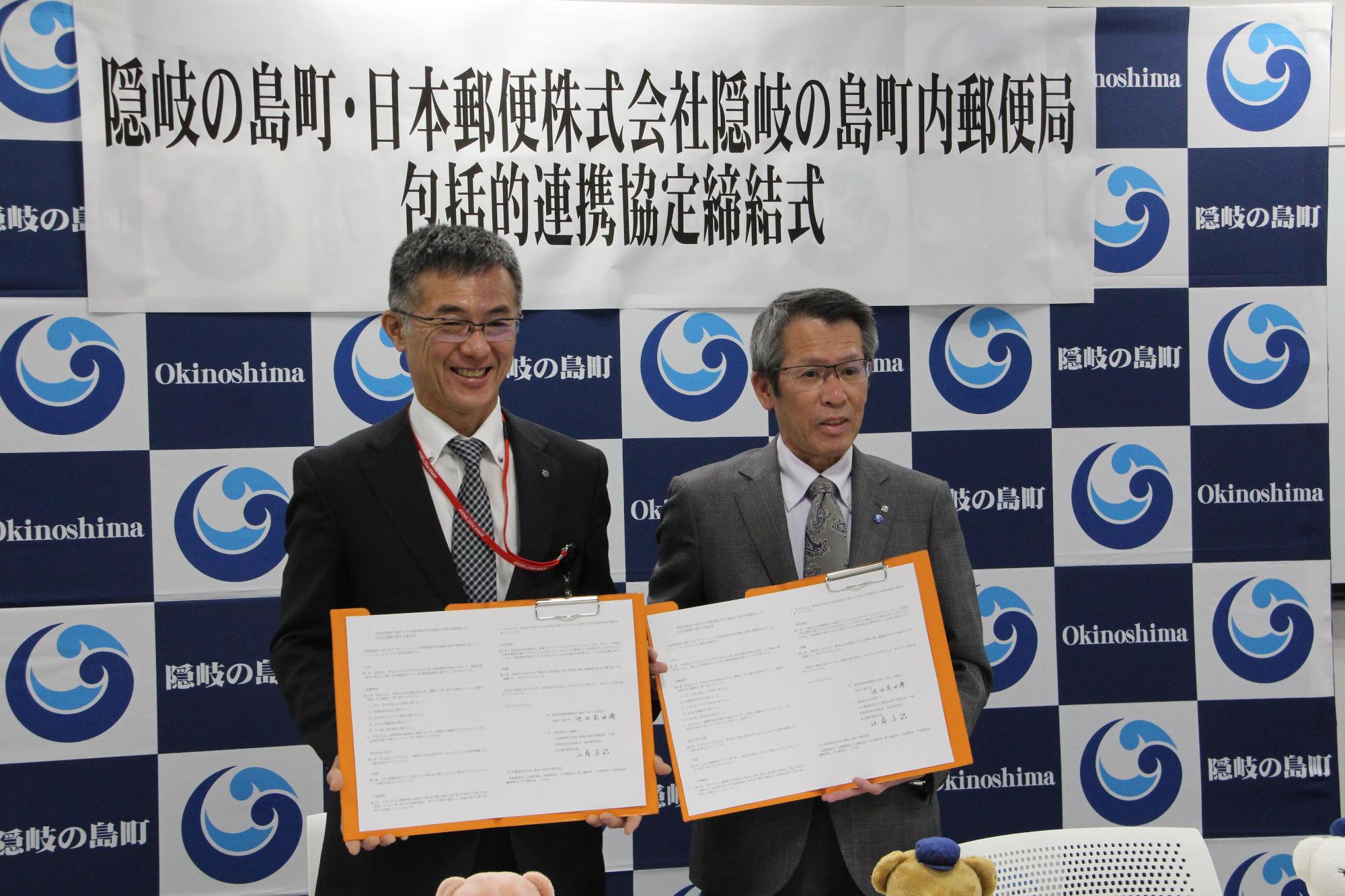 隠岐の島町・日本郵便株式会社隠岐の島町内郵便局包括的連携協定締結式と書かれた横断幕の前でスーツ姿の男性二人がそれぞれ書類を掲げている写真