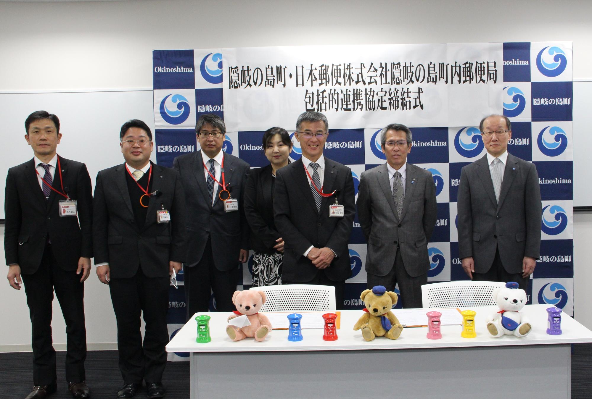 隠岐の島町・日本郵便株式会社隠岐の島町内郵便局包括的連携協定締結式と書かれた横断幕の前で一列に並んだスーツ姿の男性6名と女性一名の写真