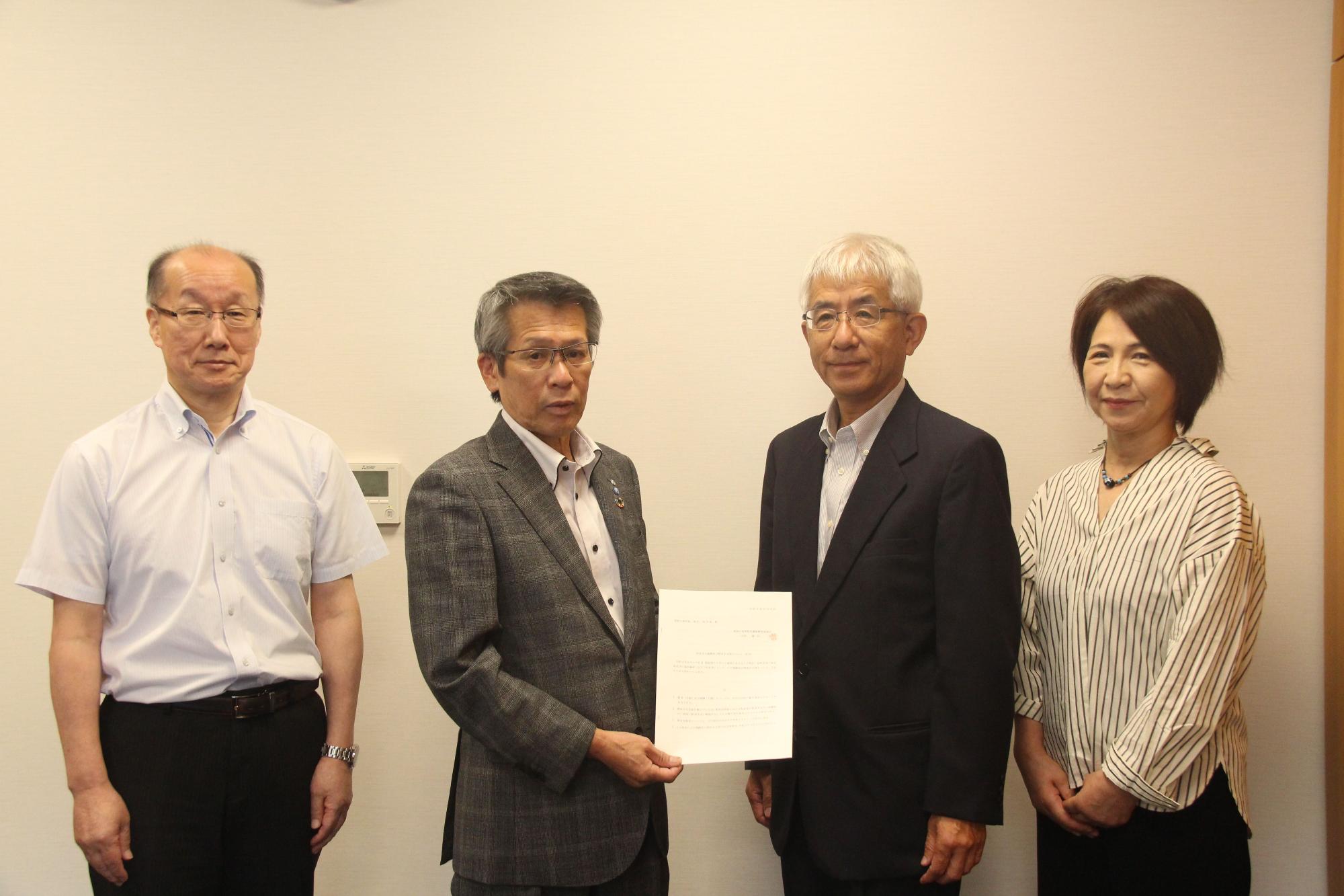 男性3人と女性1人が横一列に並んで、書面をカメラに向けている写真