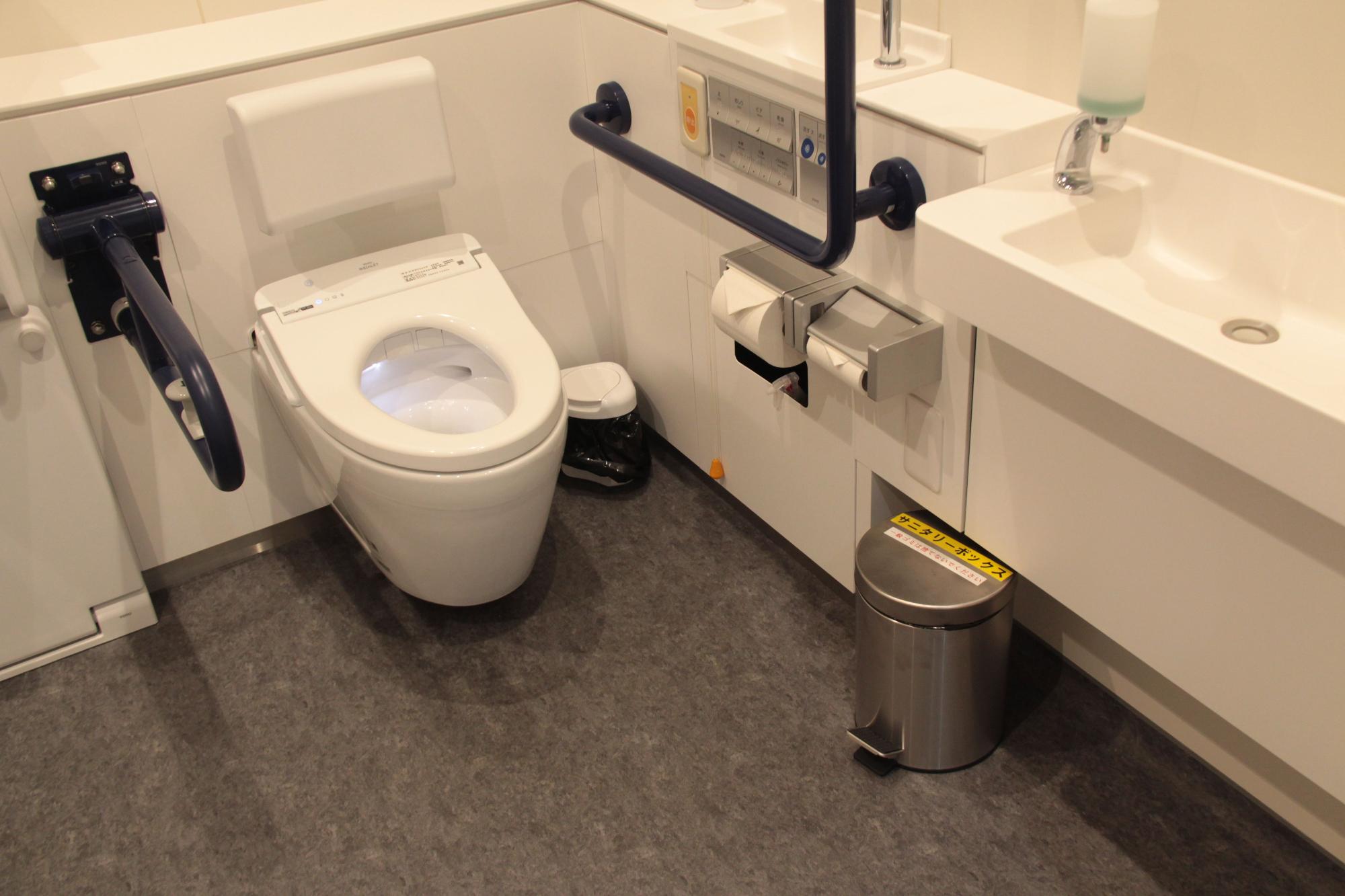 広い多目的トイレの個室内の、洗面台の下に設置された円柱型サニタリーボックスの写真