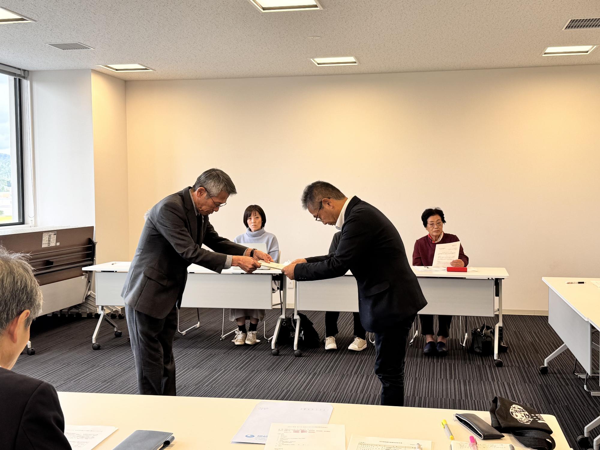 会議室にコの字型に配置された長机の席に座った複数の男女の前で二人の壮年男性が向かい合って書類を受け渡している写真