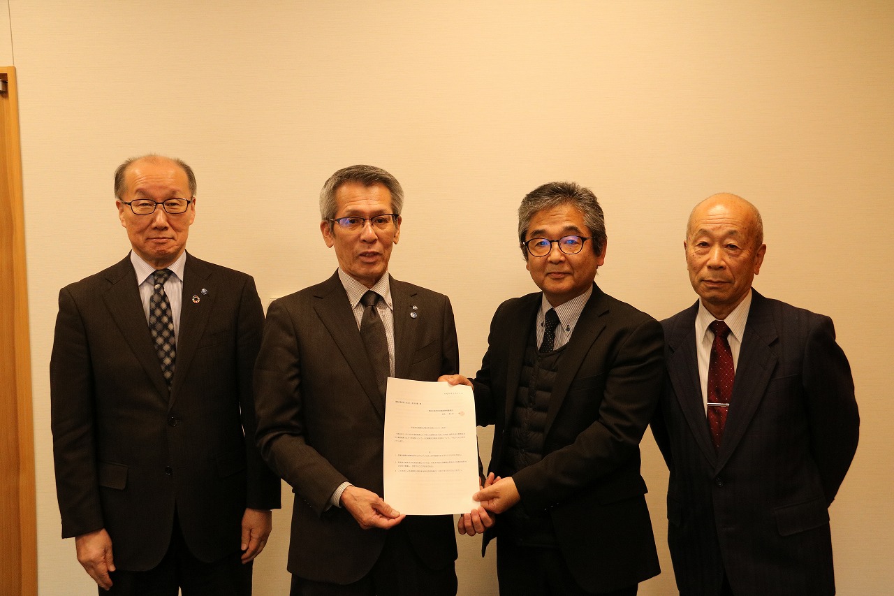 4人の男性が並んで真ん中2人が一枚の書類を掲げる写真