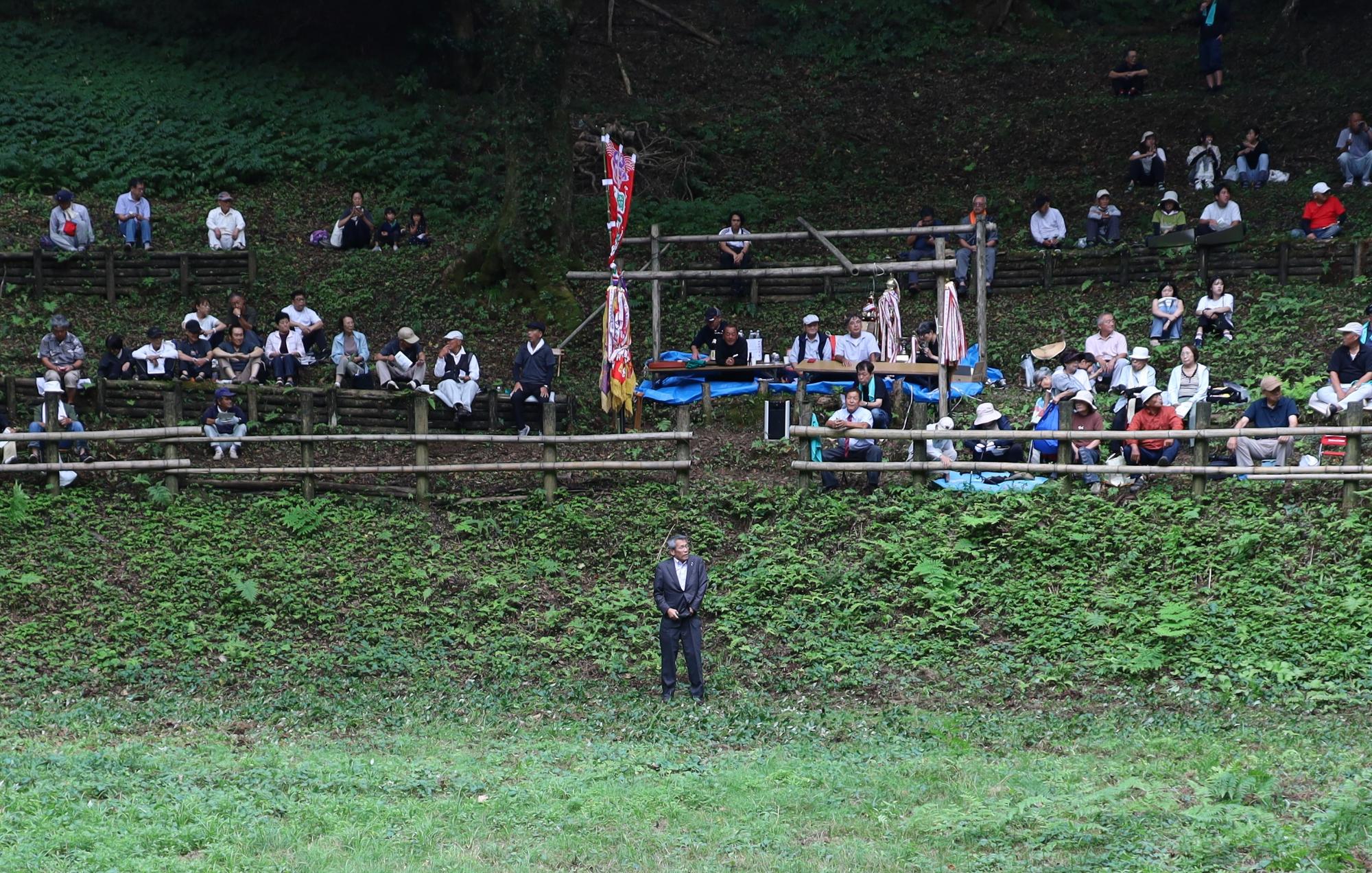 人々が腰をかけている斜面の手前でスーツ姿の男性が佇んでいる写真