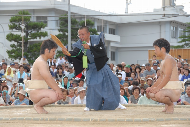 土俵の上でしゃがんで向き合う二人の褌姿の男性とその間に立つ裃を着た男性の写真