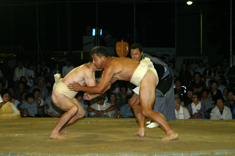 暗くなった野外の土俵で力士二人が試合をしている写真