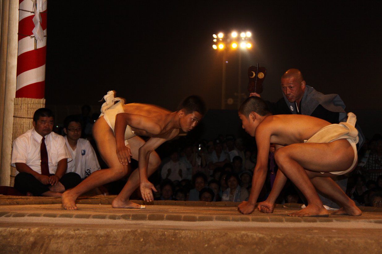 薄暗い土俵で2人の力士がお互いをにらんでぶつかりに行こうとしている写真