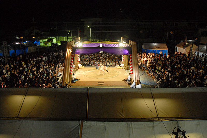 夜に屋外でライトアップされた土俵で組み合っている力士の写真