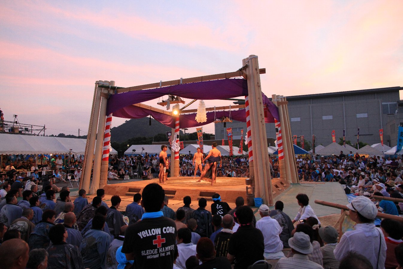 紫の幕が掛けられた屋根のない土俵に力士たちが立っている写真