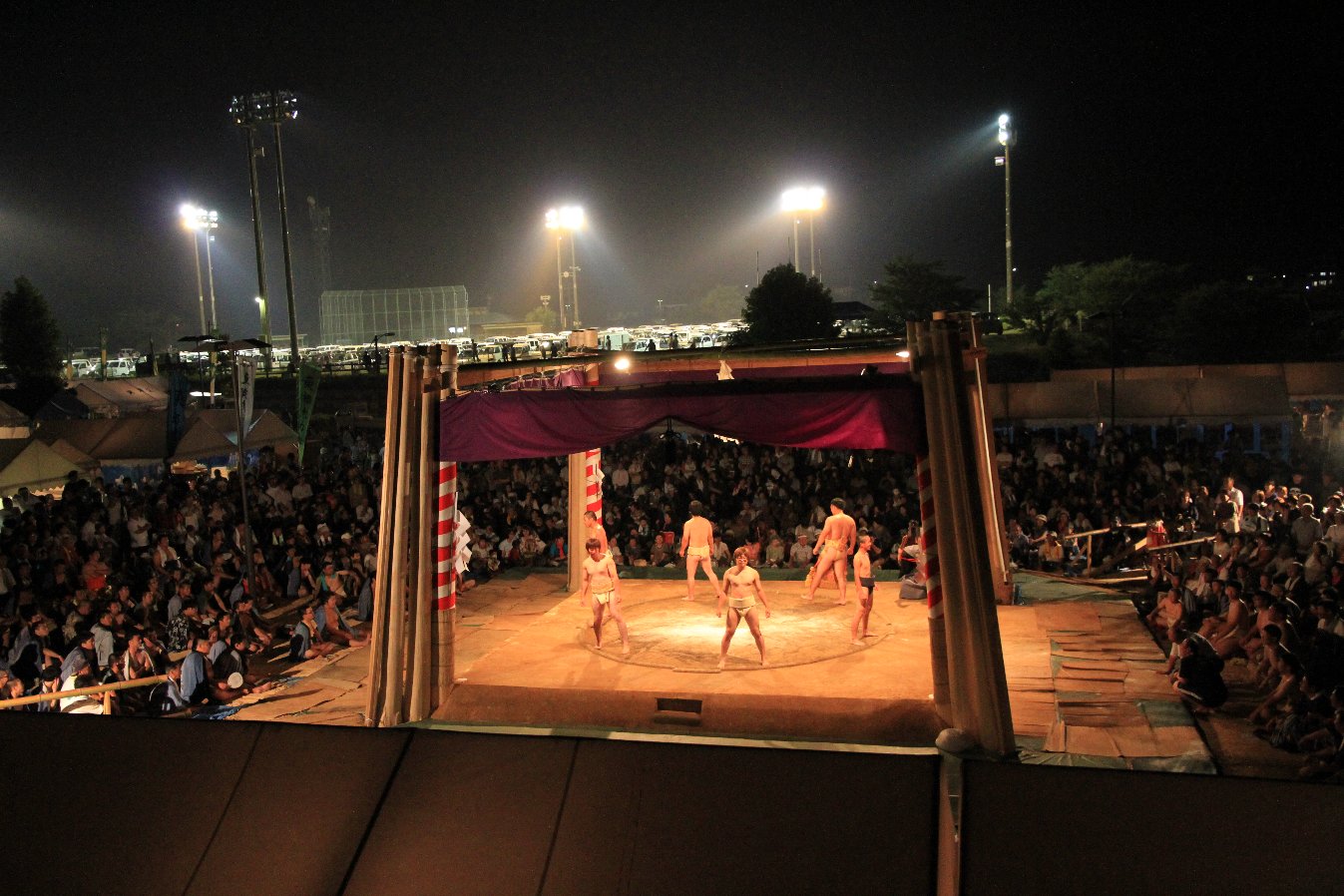 ライトに照らされた屋外の土俵で力士たちが観客を見ている写真