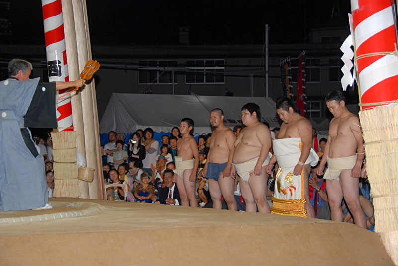 軍配扇子を掲げた行司の前で並んでいる力士たちの写真