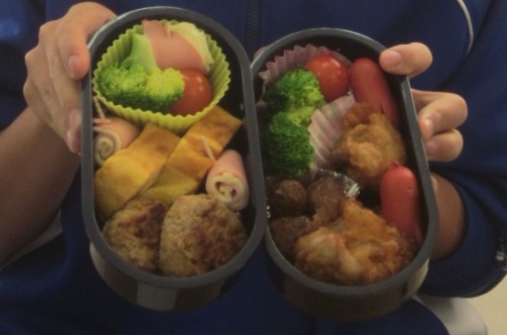 卵焼きやミートボール、野菜などが詰められた二段弁当を両手で持っている写真