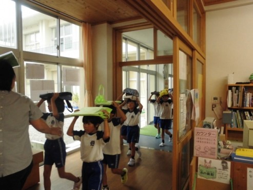 児童たちが頭の上に防災グッズを載せて避難の練習をしている写真