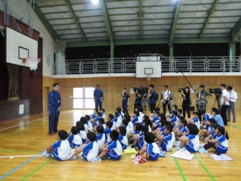 体育館で周囲にカメラや取材スタッフがいる中で児童たちが説明を聞いている写真