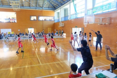 体育館で児童たちがユニフォームを着てバスケットボールの試合をし、監督が立ち上がって応援している写真