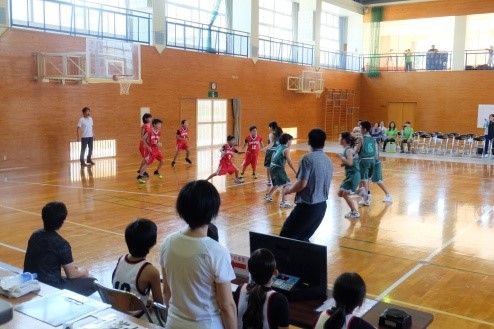体育館で児童たちがユニフォームを着てバスケットボールの試合をし、観客も試合を見ている写真