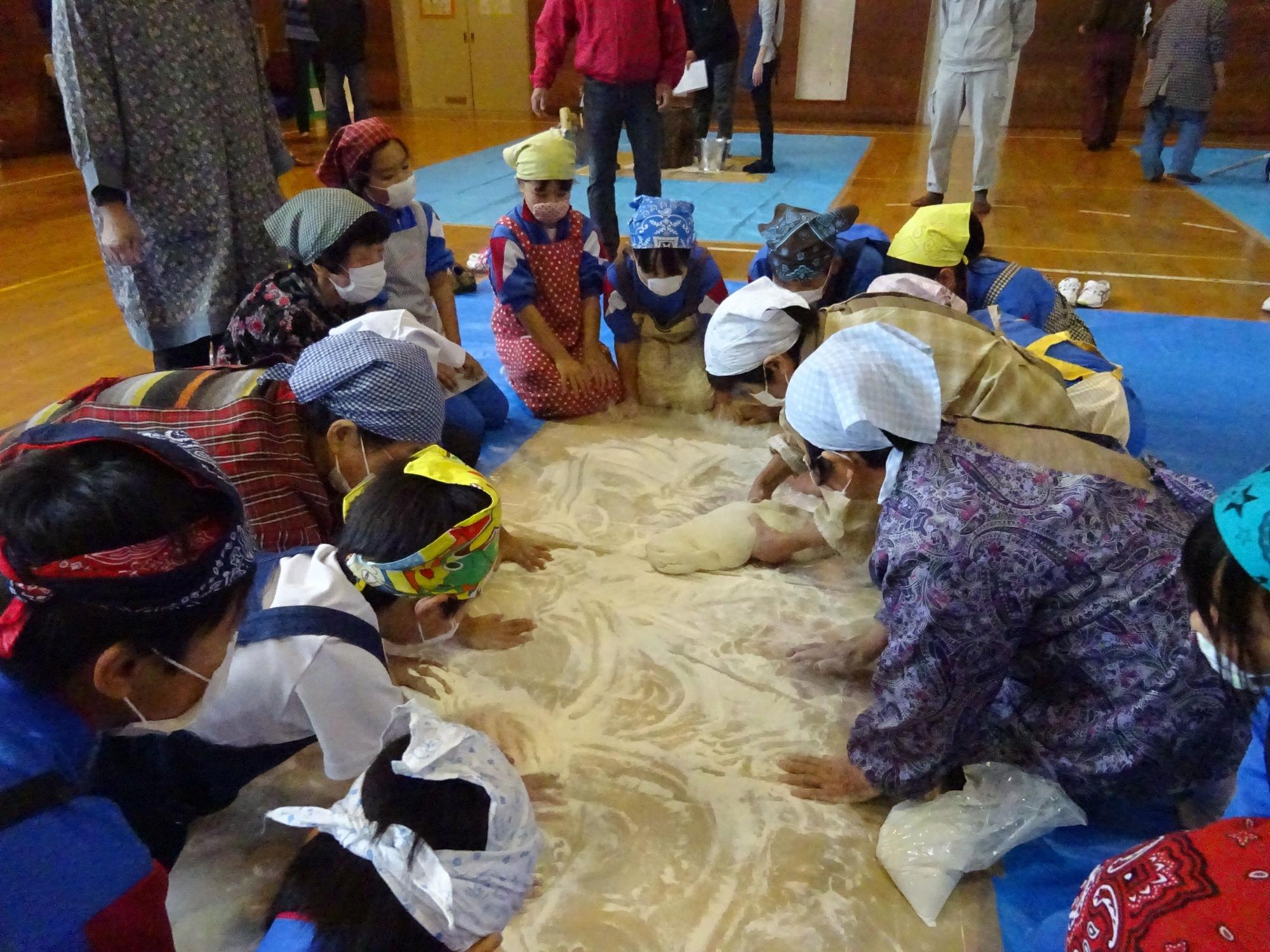 体育館で三角巾と前掛けを着用した複数の児童や大人がシートが引かれた床の上で餅をこねている写真