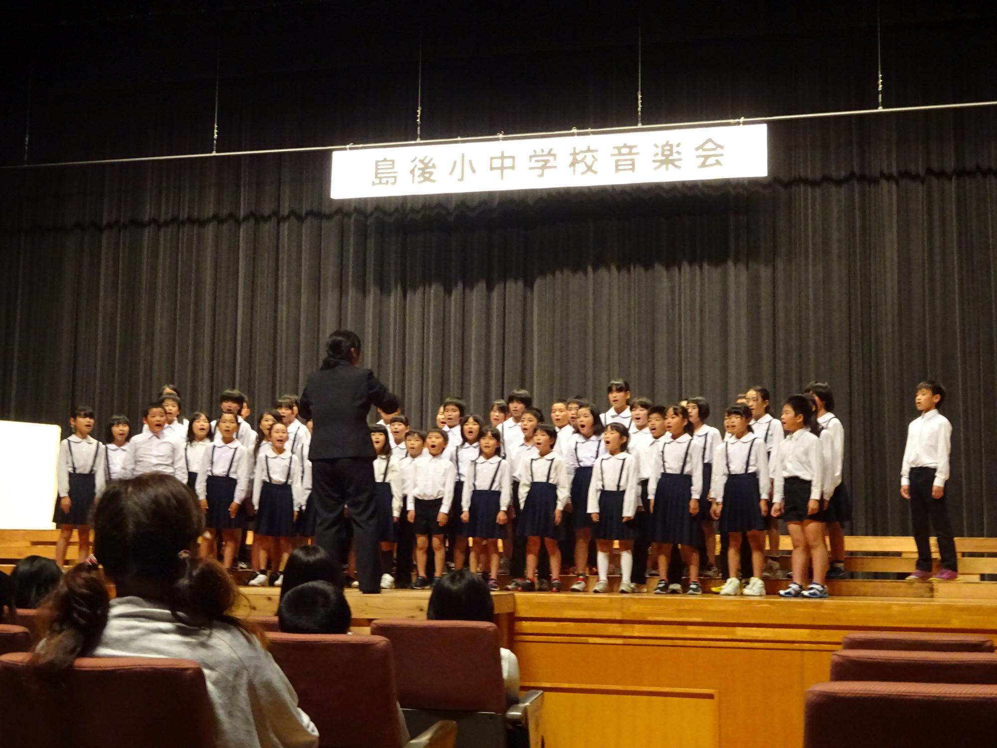 児童がステージ上で肩幅に足を広げて指揮者に合わせて合唱を行っている写真