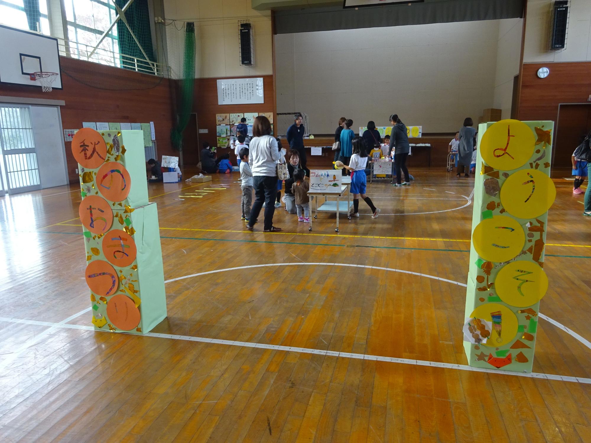 体育館で手前に「秋のおまつり」「ようこそ！」と書かれた紙製の看板の奥で児童や大人が催し物を行っている写真