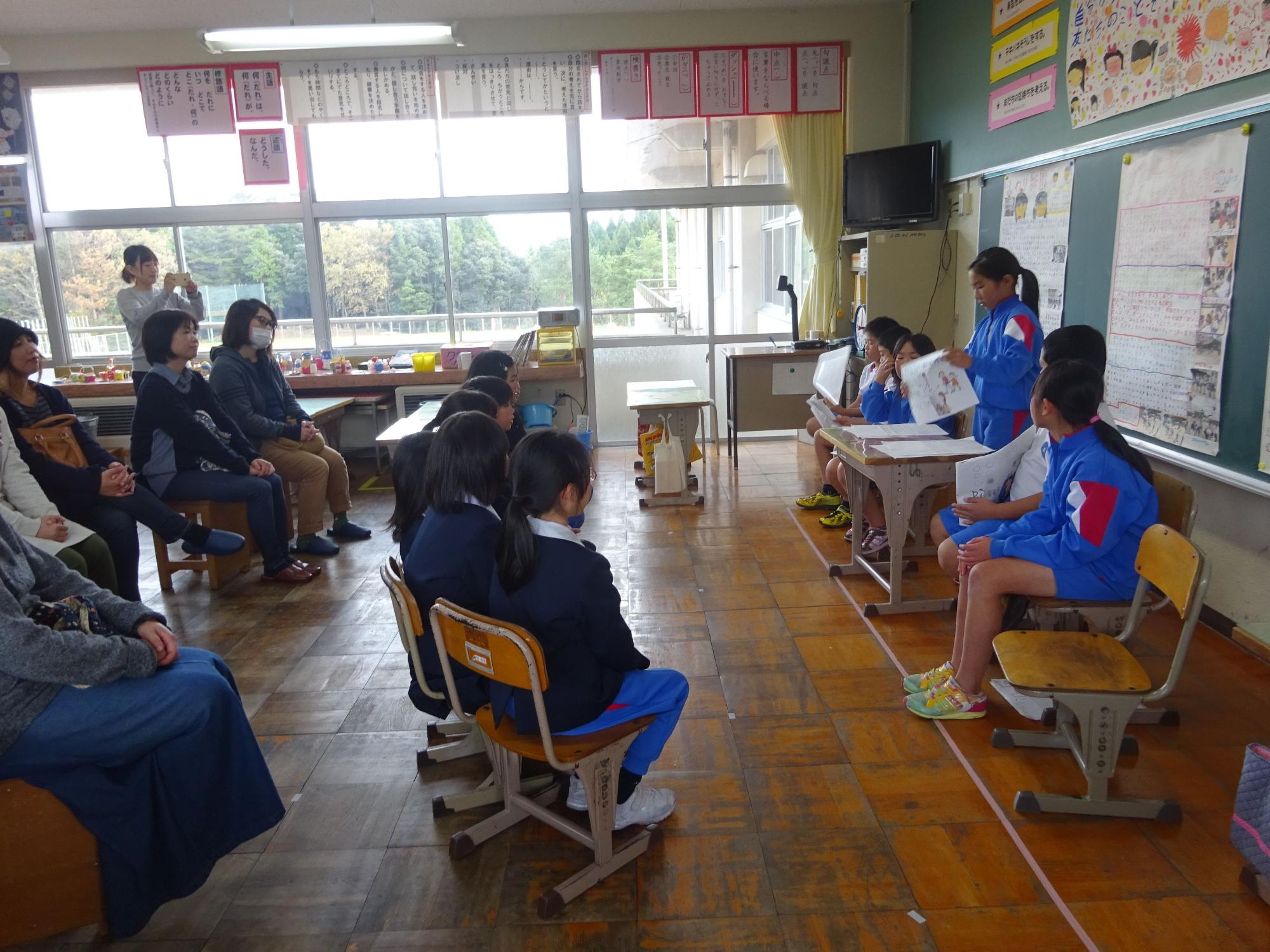 教室で児童が資料を手に持ちながら話している姿を複数の児童や大人が聞いている写真