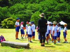公園内の草が生えた空き地で先生の話を聞いている児童たちの写真