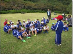 秋の遠足で芝生の上で児童たちが集まり代表の生徒の話を聞いている写真