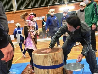 体育館で児童が杵を持ち、大人が臼に手を添えて餅つきを行っている写真