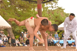 土俵の上でまわし姿の2人の児童が相撲をとっている様子を男性が見ている写真