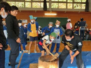 体育館で臼を手に持った児童と男性が餅つきを行っている様子を複数の人が見ている写真