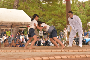 土俵の上で体操服姿の2人の児童が相撲をとっている様子を男性が見ている写真