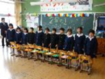 教室で横一列に並べた椅子の後ろに並んでいる児童たちの写真