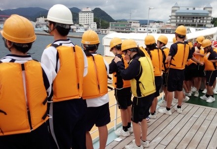 実習船で子どもたちが救命具と帽子を着け並んで海を見下ろしている写真