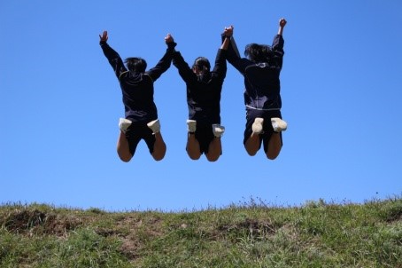 青空の中、三人の生徒が手をつなぎ地面から大きく跳び上がっている写真