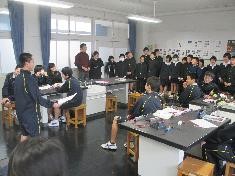 教室で多くの生徒が席に着き、一人が前に立って発表している写真