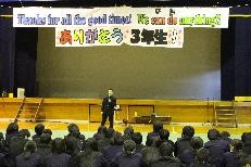 体育館で壇上の話者を生徒たちが前方で聞いている写真