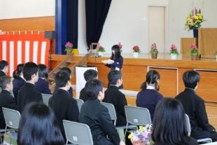 体育館の壇上で生徒が話し、客席に多くの人が座っているの写真