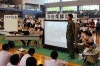 体育館で制服の自衛隊員が生徒に説明をしている写真