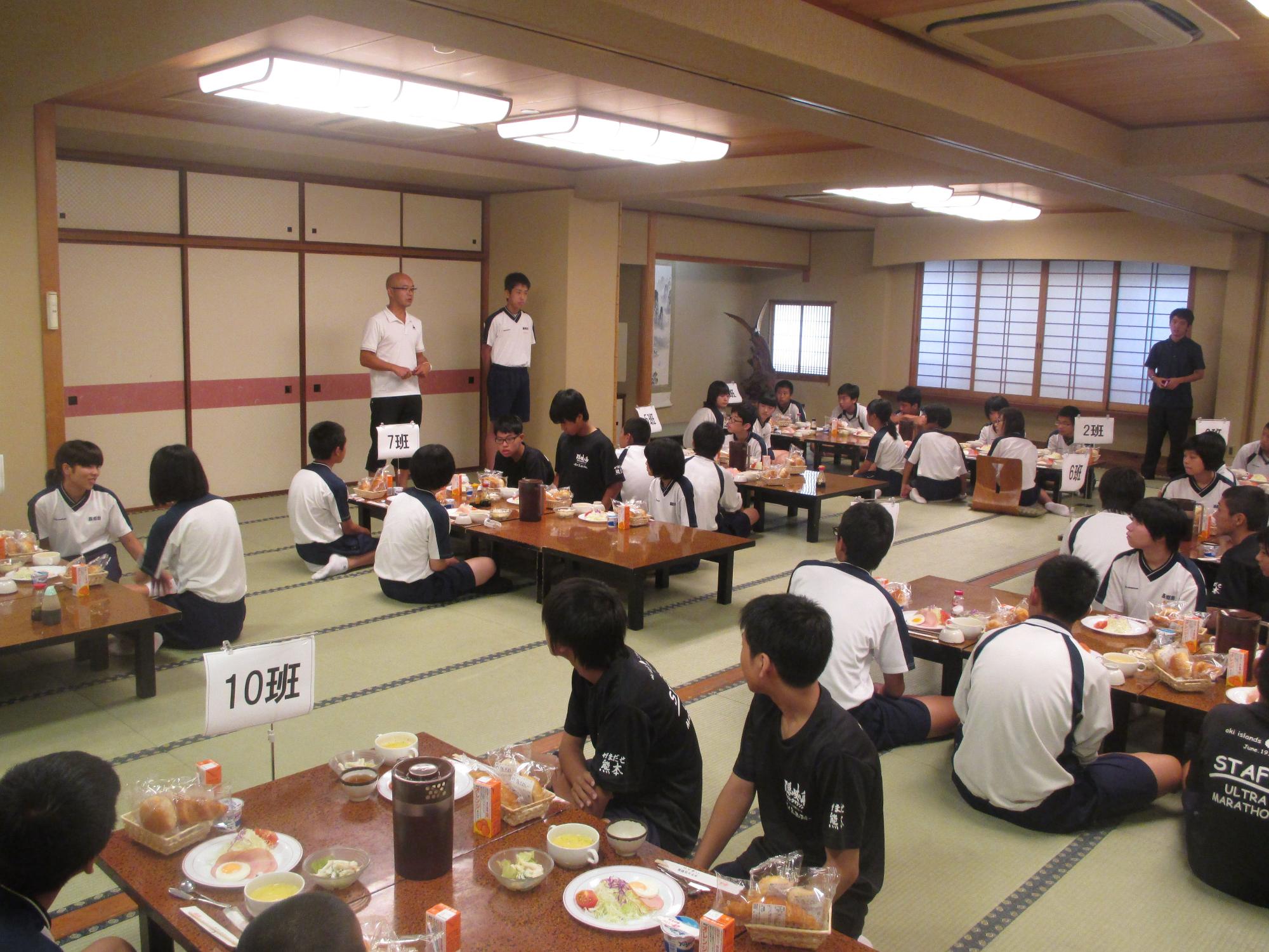 広い和室で体操服姿の生徒が班ごとに座り、配膳された食事を囲んで前方に立っている大人の話を聞いている写真