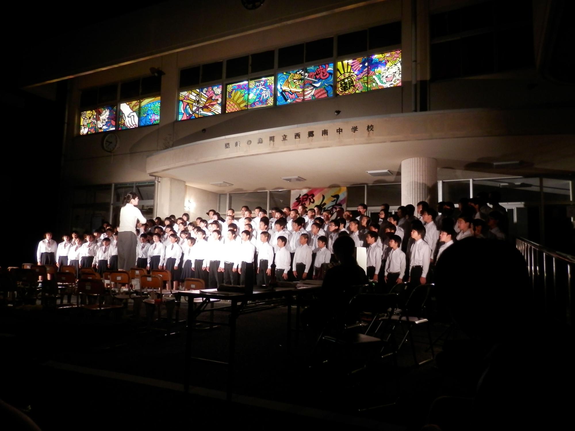 校舎前で多くの生徒が指揮者の方を向き合唱している様子をスポットライトで照らされた写真