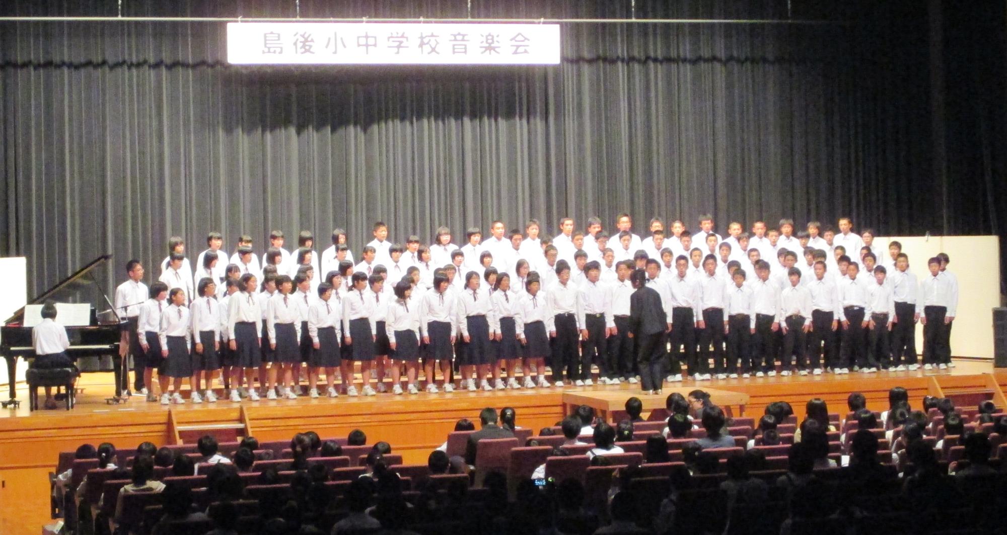 ステージ上に大勢の生徒が指揮者の方を向きながら合唱している様子を複数の人が着席に座り見守っている写真