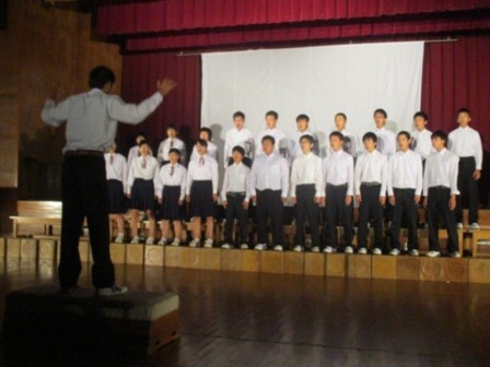 体育館で男女の生徒たちが指揮に合わせて合唱している写真