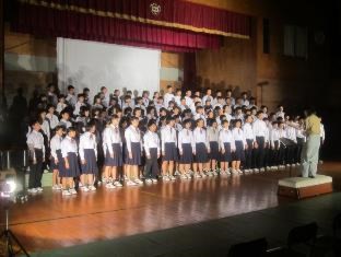 多数の生徒が体育館の舞台上に並び、指揮者の合図で合唱している写真