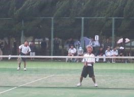 屋外のテニスコートで生徒がテニスのダブルスをしている写真