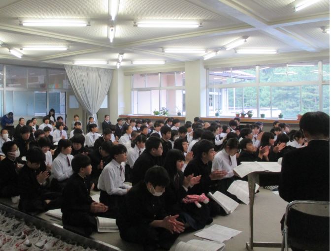 床に座って資料を見ながら話を聞いて拍手している生徒たちの写真