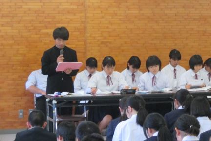 制服を着た生徒がマイクを使って説明をし、周りの生徒は資料を見ながら聞いている写真