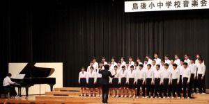 ステージ上でピアノを伴奏している生徒の横に複数の生徒が指揮者に合わせて合唱をしている写真